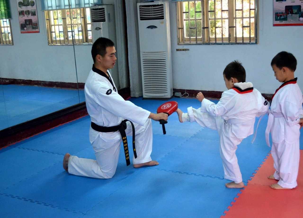 Taekwondo isntructor holding kicking bag as 2 young male students stand in line to practise their kicks