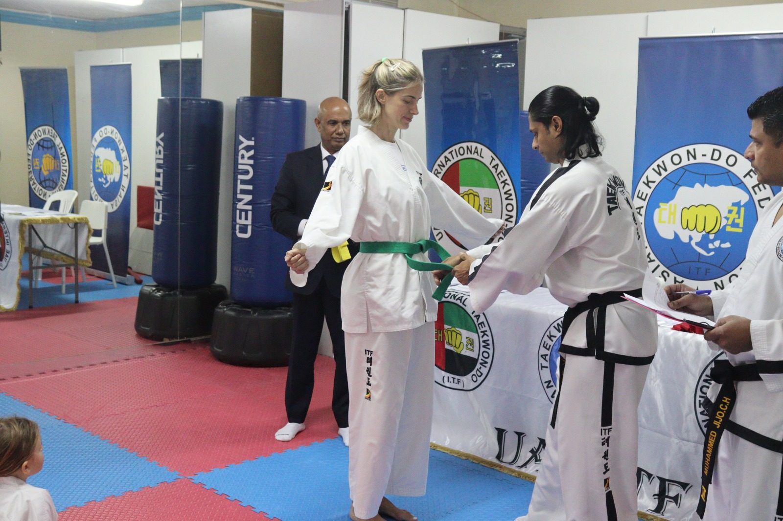 Yound woman wearing taekwondo uniform getting a green belt tied around her waist by instructor