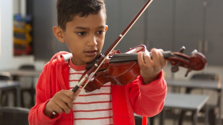 Front view of cute mixed-race schoolboy playing violin in classroom at elementary school