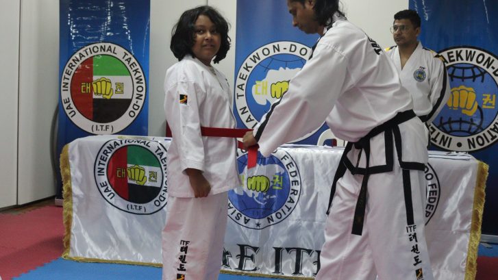 Male student wearing taekwondo uniform with a red belt being tied around his waist by instructor