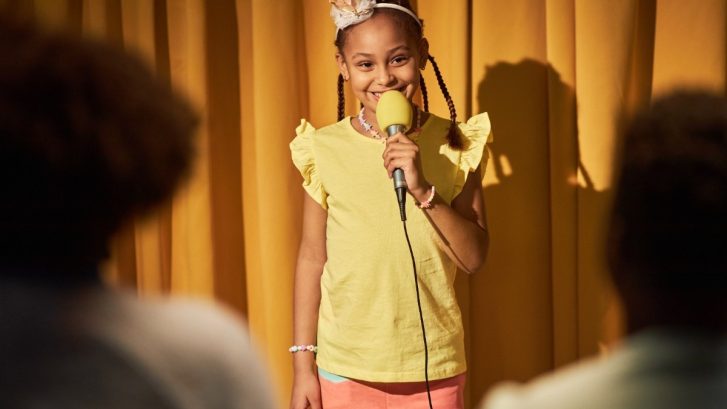 Smiling Girl Speaking To Microphone Performing for Parents