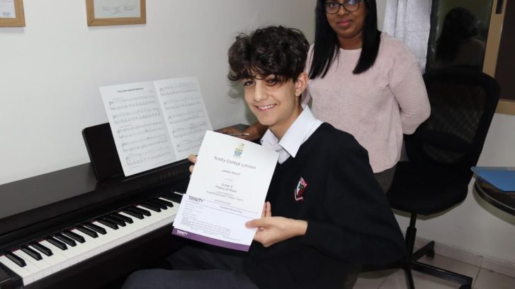 A young boy showing off his certificate as he sits in front of the piano with his instructor behind him
