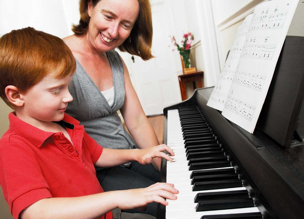 A little boy and his mom smiling as she teaches him how to play piano